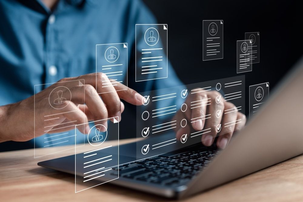 Close-up of hands operating a laptop with digital employee profiles and checklists projected on the screen, representing an HRIS system in action.
