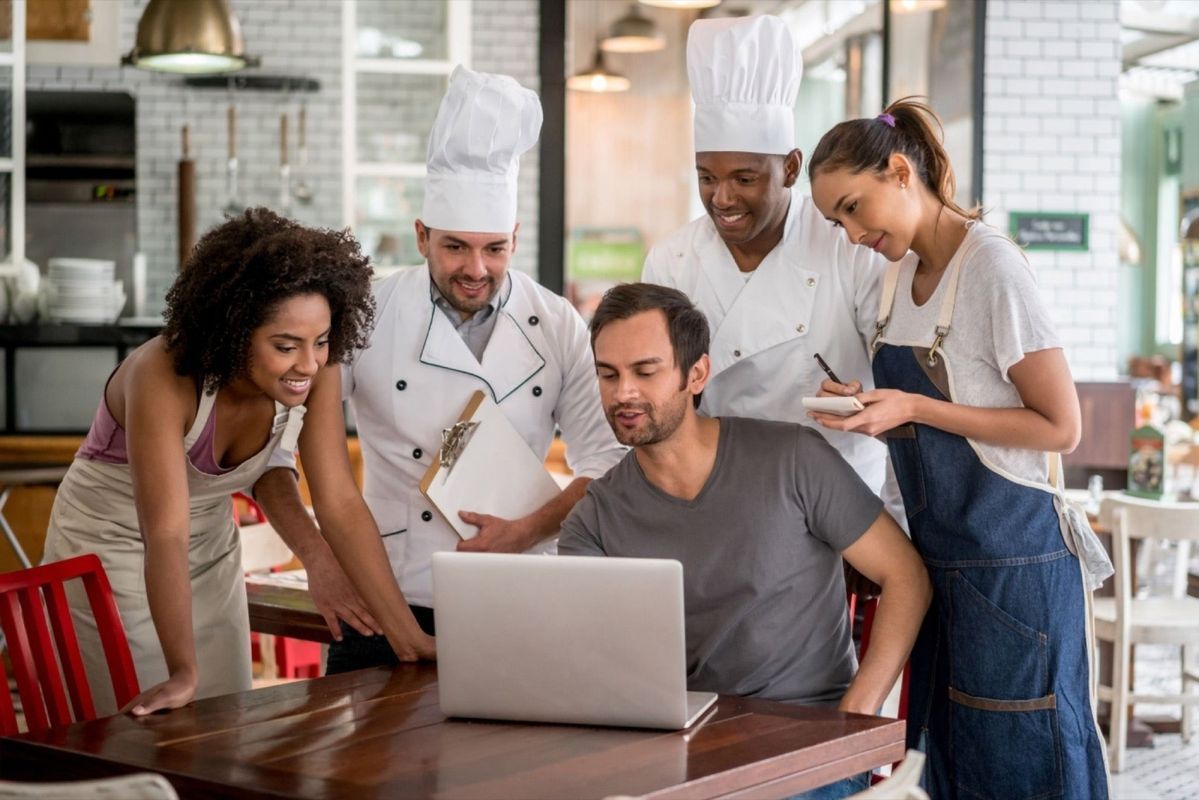 Restaurant staff and managers gathered around a laptop, reviewing shift schedules and HR processes using a digital HRIS platform.