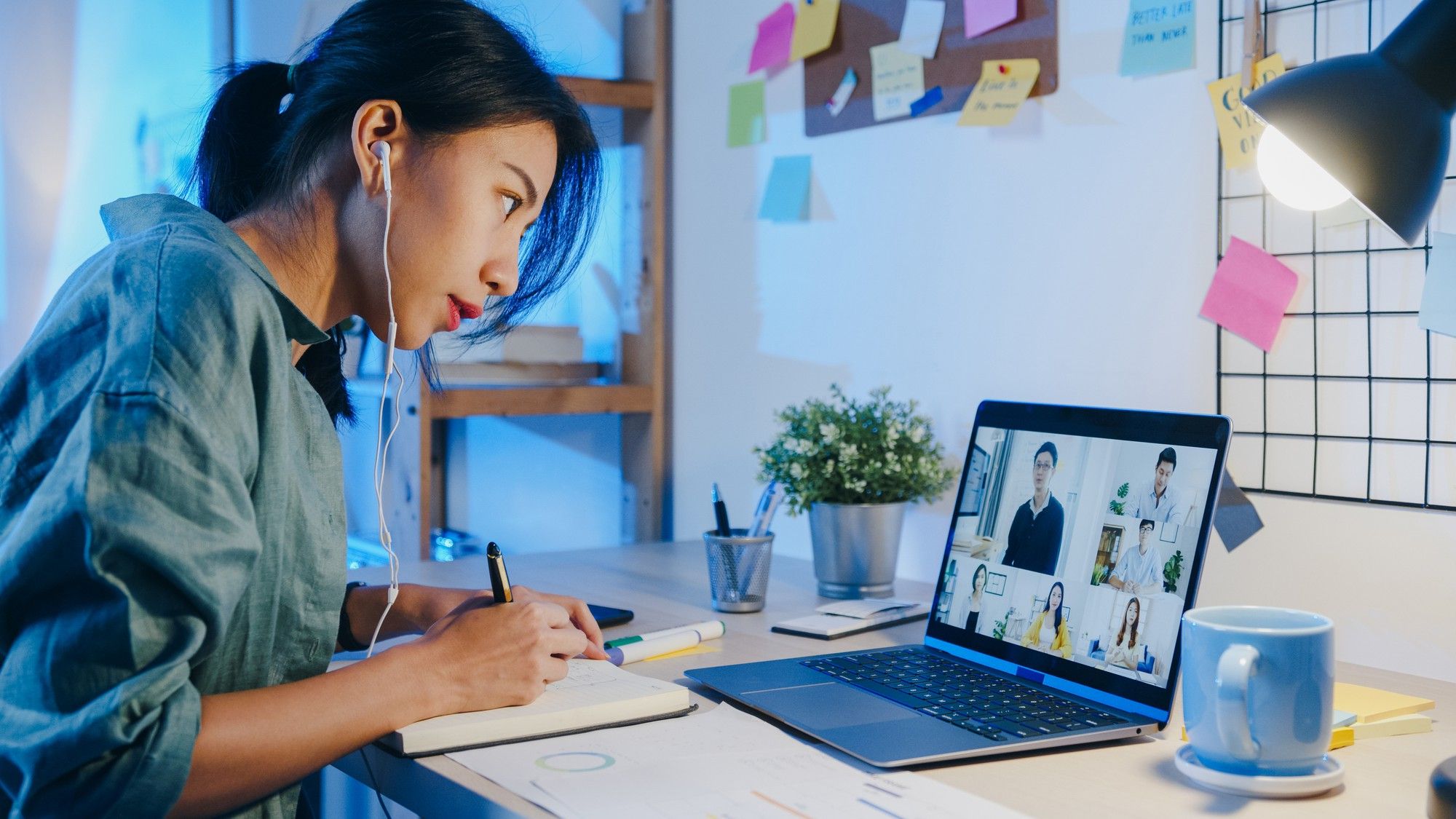 A woman taking notes while attending an online video conference class on her laptop, representing modern e-learning and EdTech solutions.