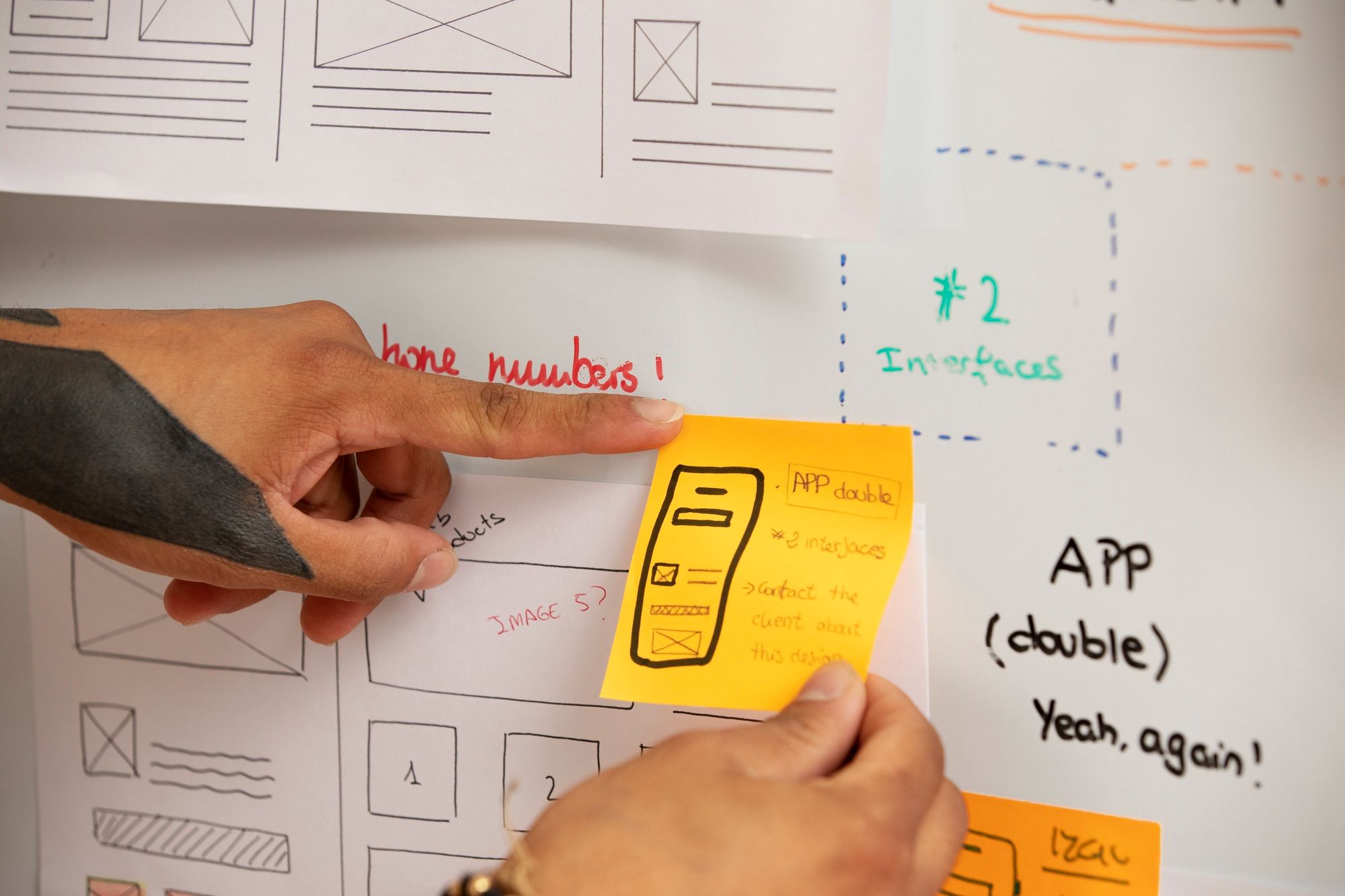 A person pointing at a sticky note with an app interface sketch on a planning board, illustrating UI/UX design workflows supported by Figma plugins for faster EdTech or school app development.